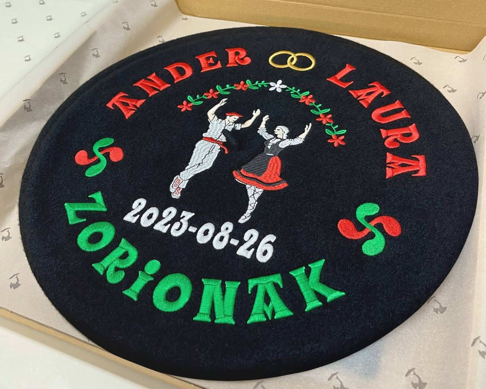 Txapela negra personalizada para boda en su caja. Bordada con nombre de los novios, dantzaris, lauburus, alianzas y lazo floral.