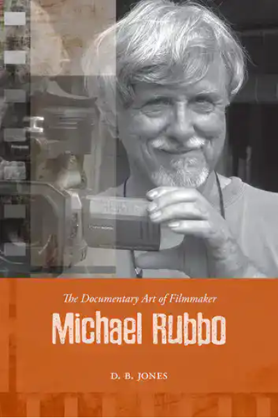 Photograph of Michael Rubbo holding a camera, smiling, in a documentary filmmaking setting.