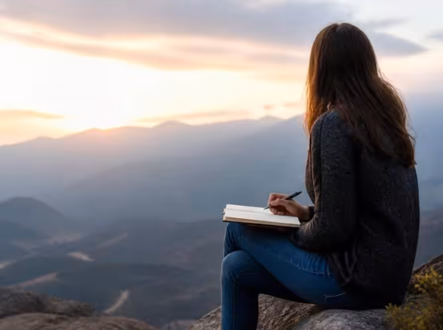 Woman sitting on rock reading a book with mountain view at sunset.
