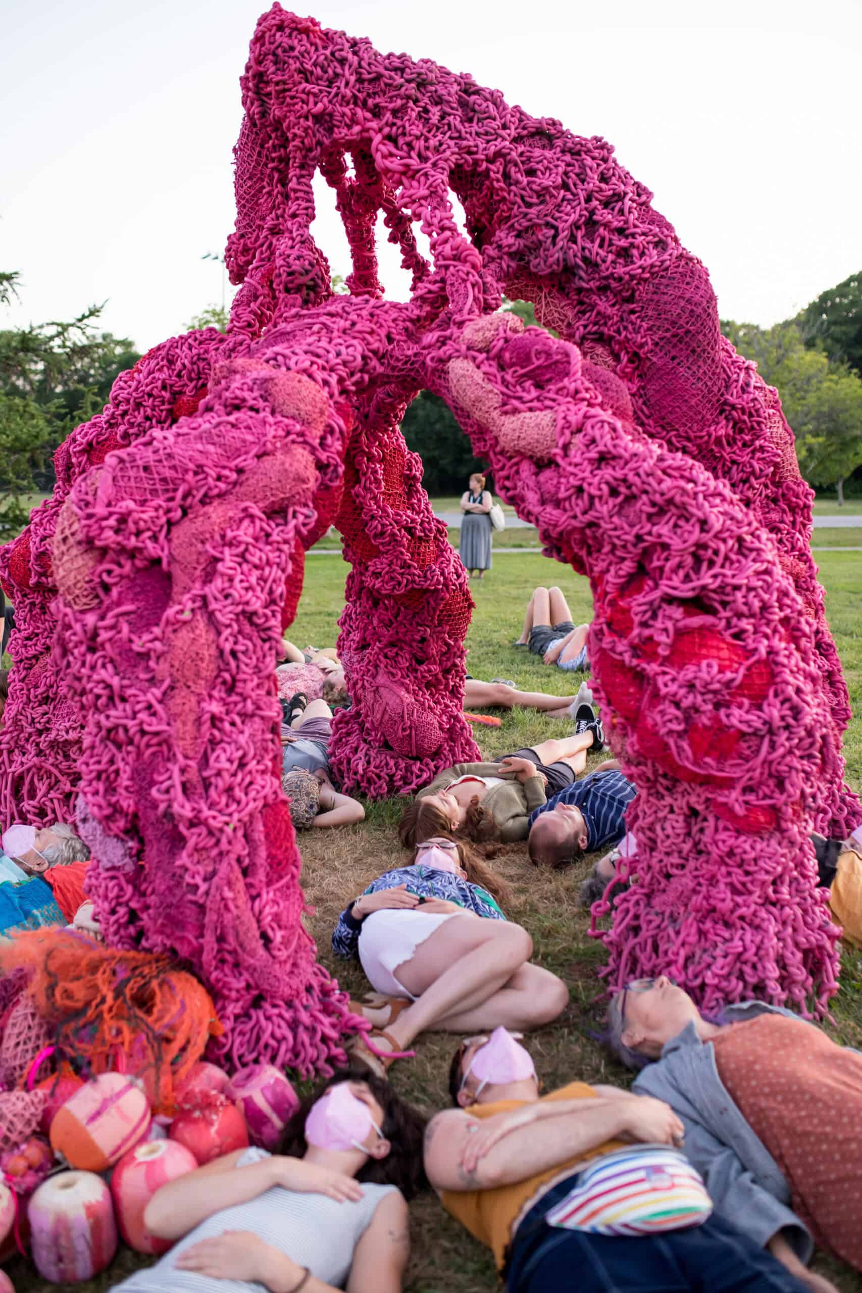 Large pink floral sculpture arching over people lying on the ground at an outdoor event.