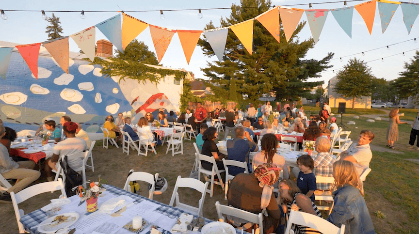 Outdoor community event with tables, colorful banners, and people celebrating together.