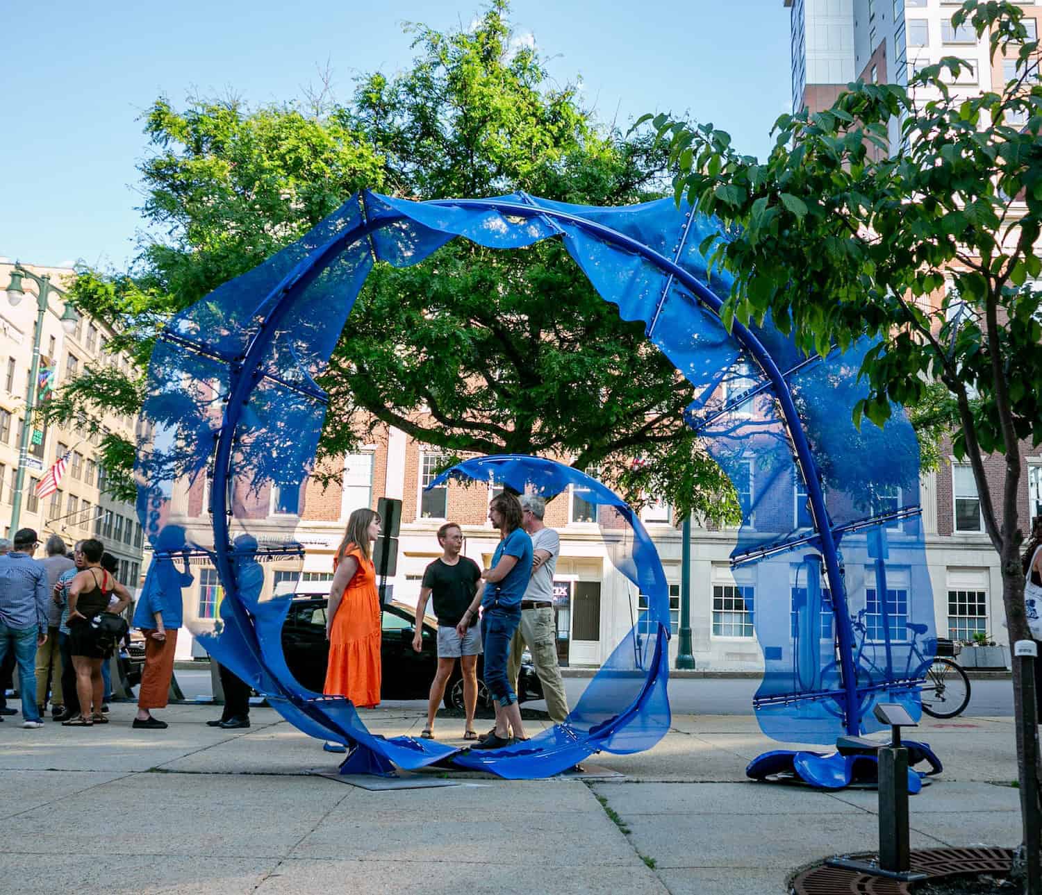 A large blue fabric art installation in a city park, with people interacting around it.