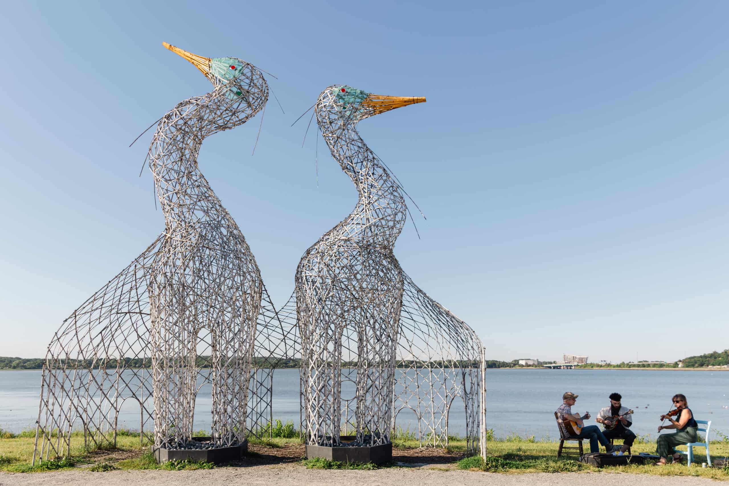 Large metal heron sculptures by TEMPOart near a river with people sitting nearby.