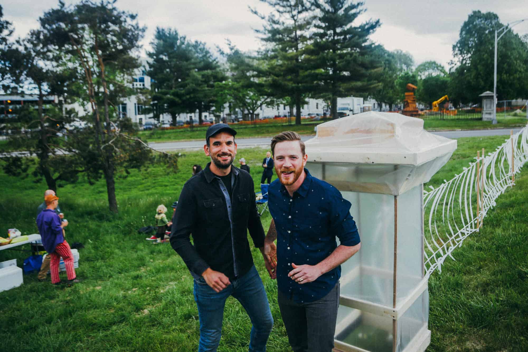 Two men standing outdoors near a large white art piece in a park setting.