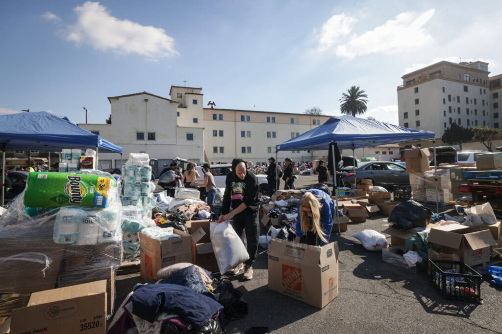 Cluttered outdoor storage area with people sorting and packing items under blue canopies, in a parking lot.