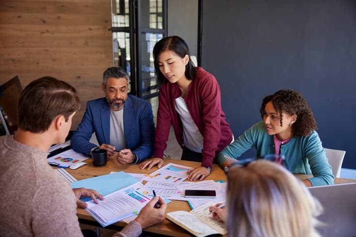 A group of professional business people discussing project details around the conference table. Sales team members reviewing documents and sharing insights on profit and loss. Asian businesswoman showing business documents during a brainstorming in creative office.