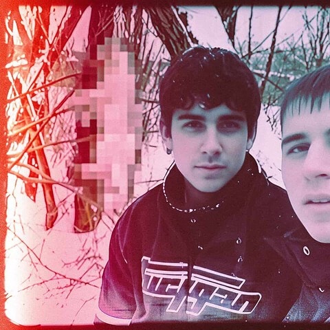 Two young men in a dark, eerie forest with a pixelated object in the background, reflecting human darkness and unsettling themes.
