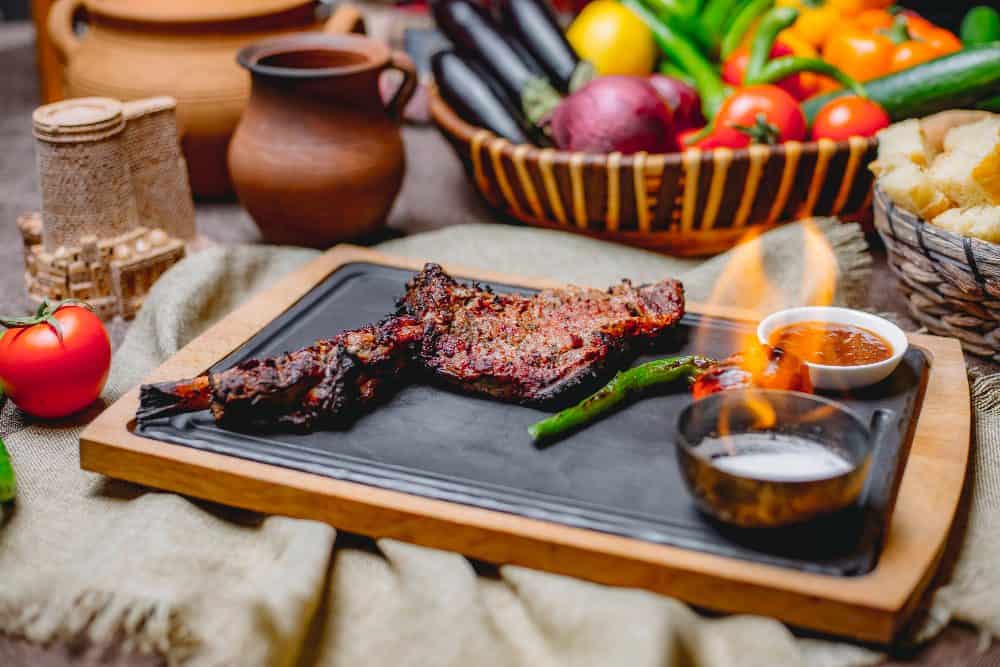 A close-up of a grilled steak or lamb leg on a black platter, served with a grilled green pepper and two small bowls of sauce and seasoning.