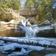 Snow-covered waterfall in a forested area, winter landscape, cold weather scene, frozen water, scenic outdoor nature.