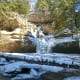 Snow-covered waterfall in a forested area, winter landscape, cold weather scene, frozen water, scenic outdoor nature.