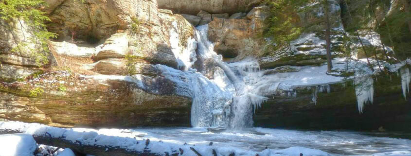 Snow-covered waterfall in a forested area, winter landscape, cold weather scene, frozen water, scenic outdoor nature.