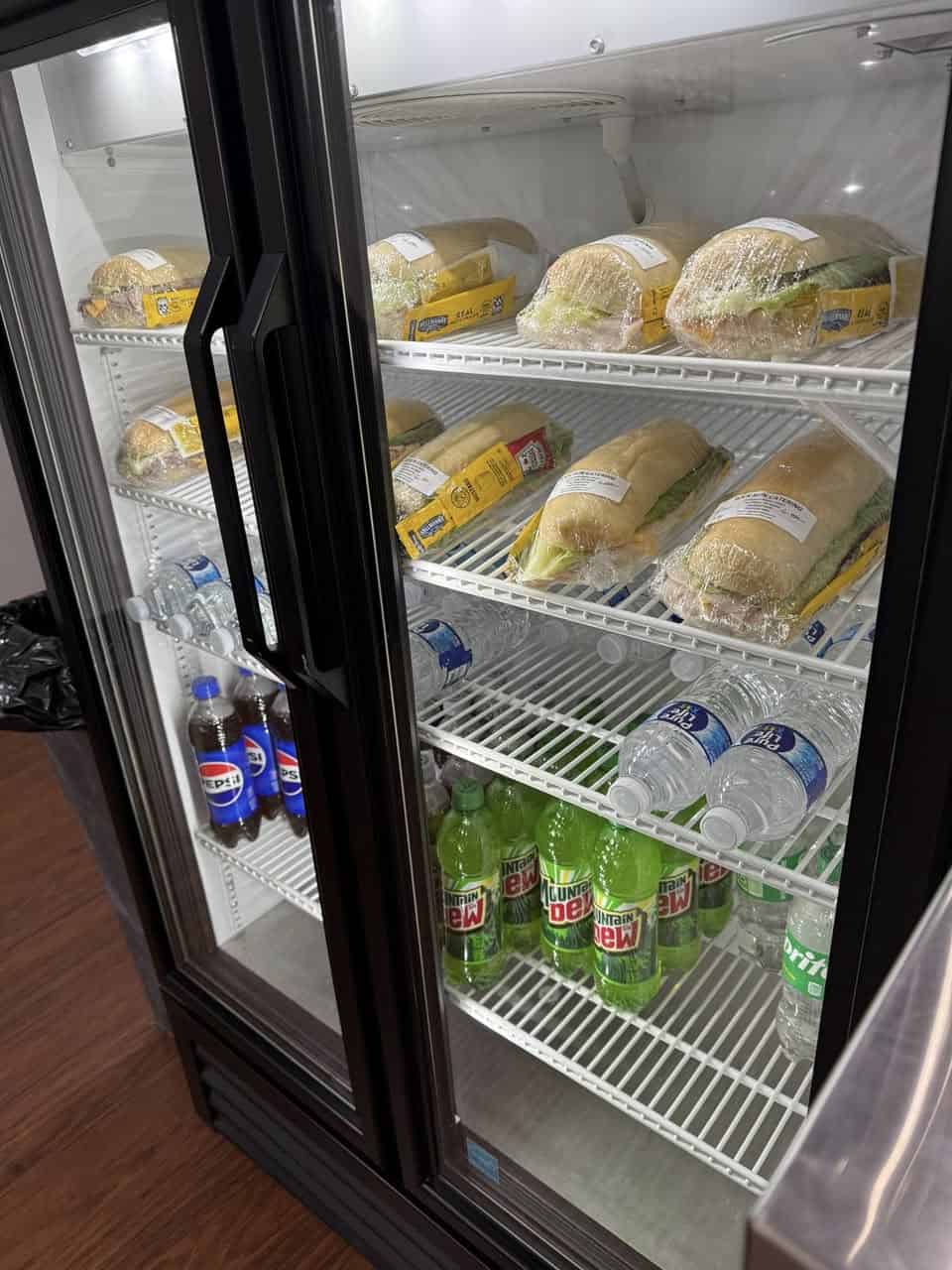 Deli sandwiches in refrigerated display, bottled water, soda, and Mountain Dew drinks, convenient for quick grab-and-go meals.