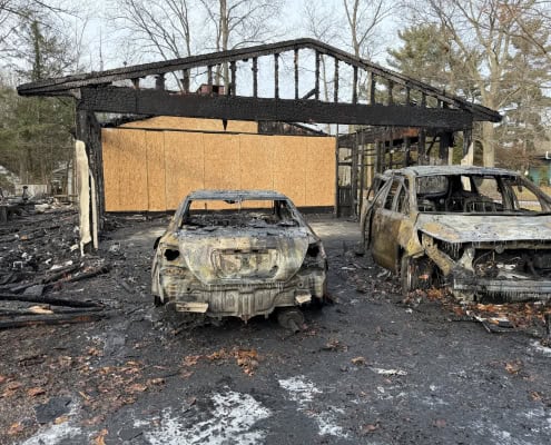 Charred and destroyed cars in front of a burned-down house following an intense fire.