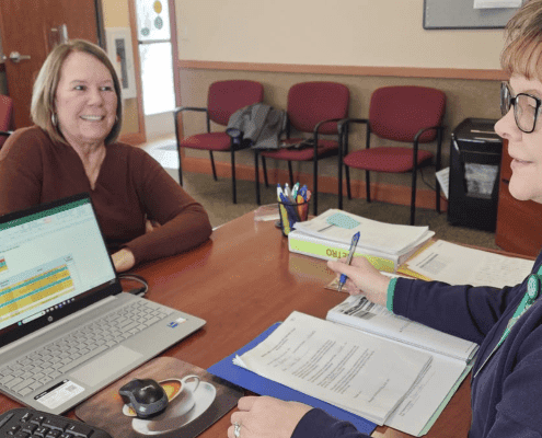 A woman and a woman sit at a table with documents, a laptop, and office supplies, engaging in a professional discussion.