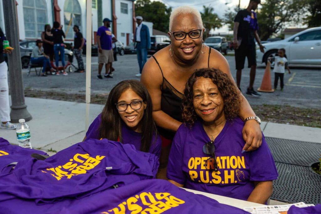 Three women smiling and posing together at an outdoor event. The two seated women wear purple "Operation P.U.S.H." shirts, with the elder woman also wearing sunglasses. A younger woman stands behind them, wearing a black shirt. Merchandise is displayed on a table.