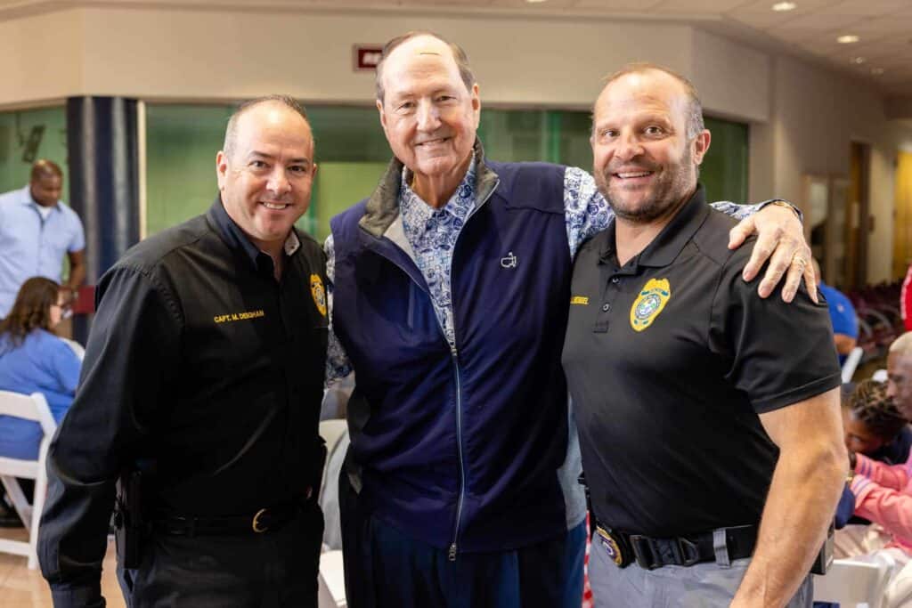 Three men are standing together and smiling indoors. The man in the center, wearing a blue jacket, is being hugged on each side by the other two men, who are dressed in black uniforms. They appear to be posing for a group photo at a casual event.