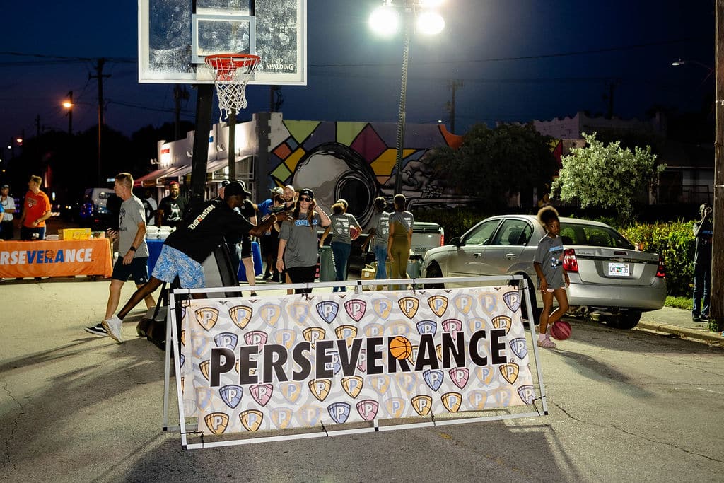 A group of people play basketball outdoors at night. The scene is illuminated by overhead lights, and a banner with the word "PERSEVERANCE" is displayed prominently in the foreground. A mural of a face is painted on a wall in the background.