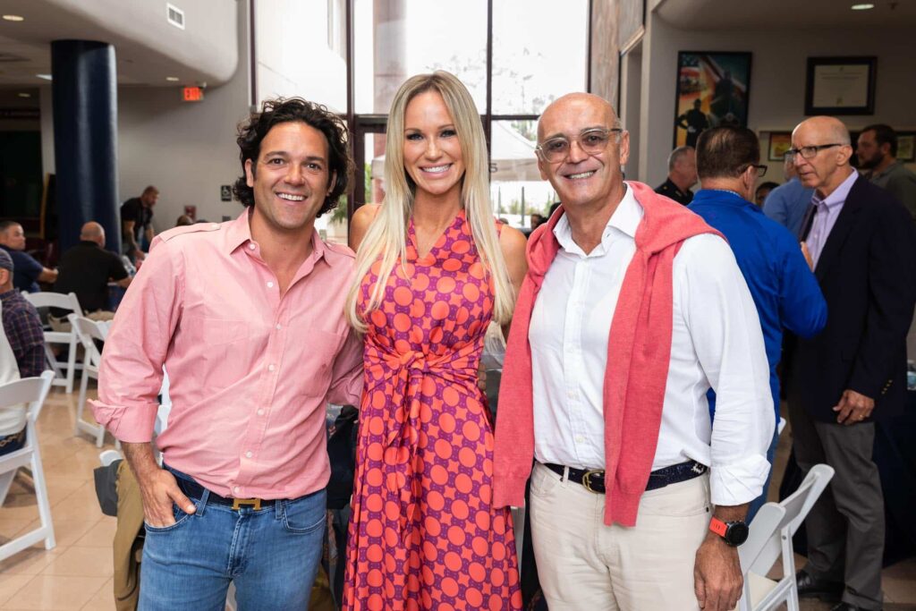 Three people are smiling and posing together indoors at an event. The person on the left wears a light pink shirt and jeans, the person in the middle wears a pink and purple patterned dress, and the person on the right wears a white shirt, beige pants, and has a pink sweater draped over their shoulders.