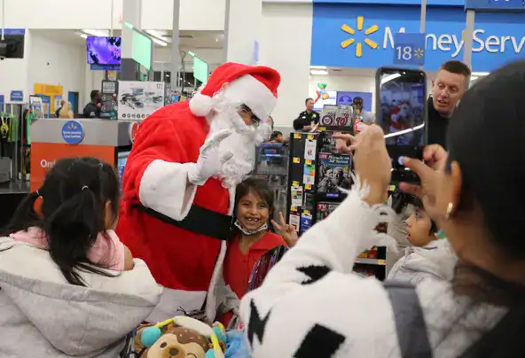 A person dressed as Santa Claus poses with a group of children in a store. One child stands close to Santa, smiling for photos taken by adults with smartphones. The background includes store signs and a Christmas toy in a shopping cart.