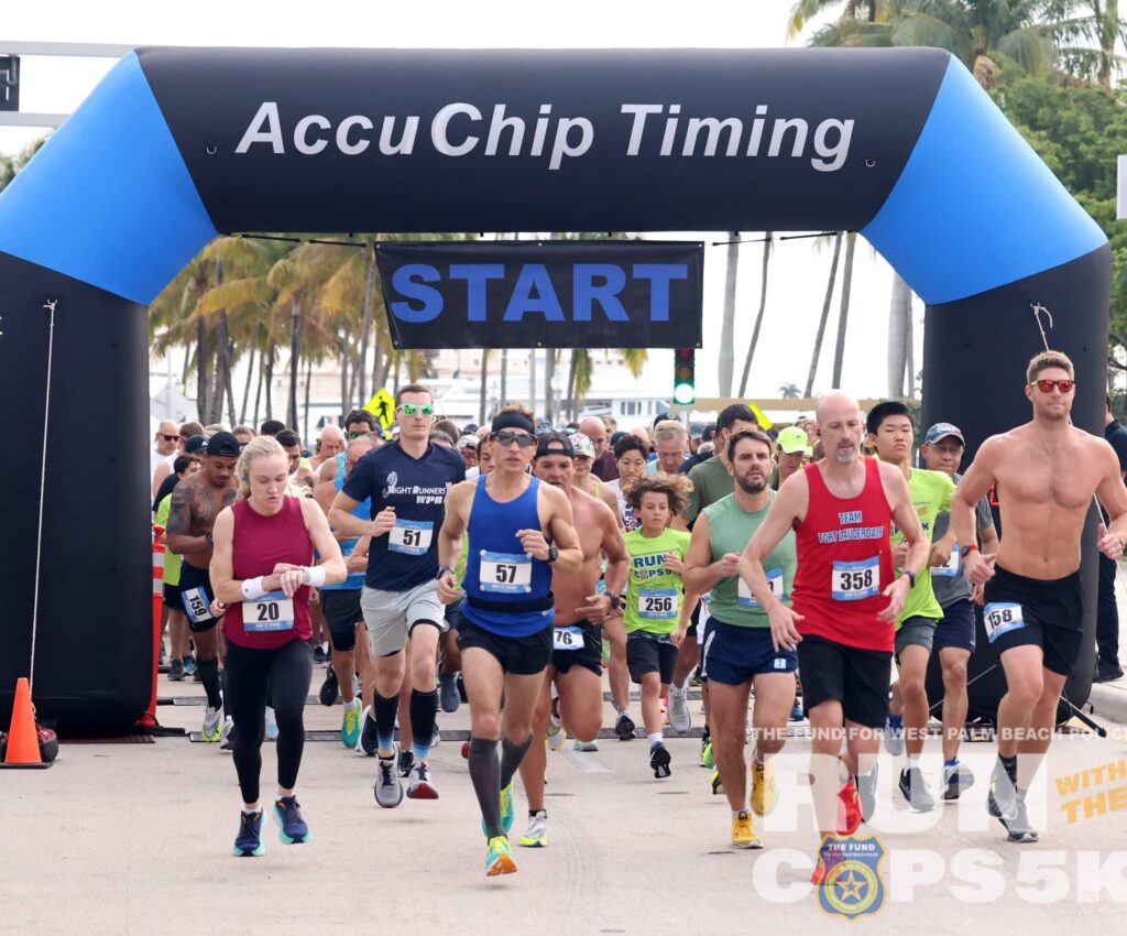 A large group of runners beginning a race under an inflatable arch labeled "AccuChip Timing" with the word "START" displayed on a banner. The participants wear various athletic attire and numbered bibs. Palm trees and clear skies are visible in the background.