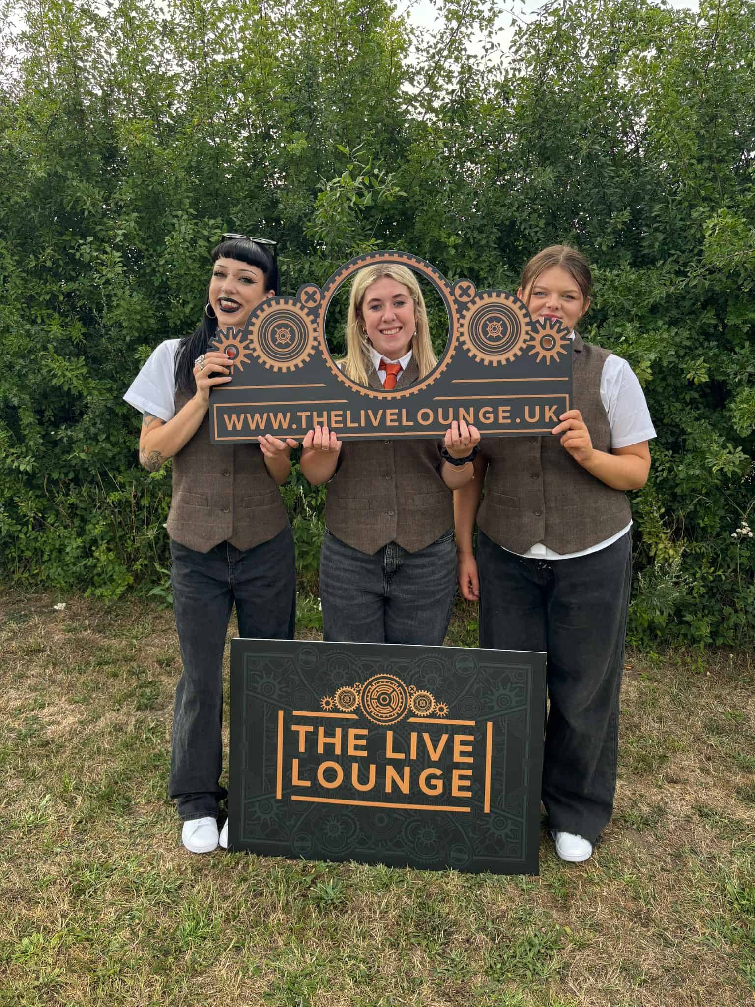 Cheerful group of women holding a promotional sign for The Live Lounge, showcasing a fun, lively atmosphere for live performances and events.