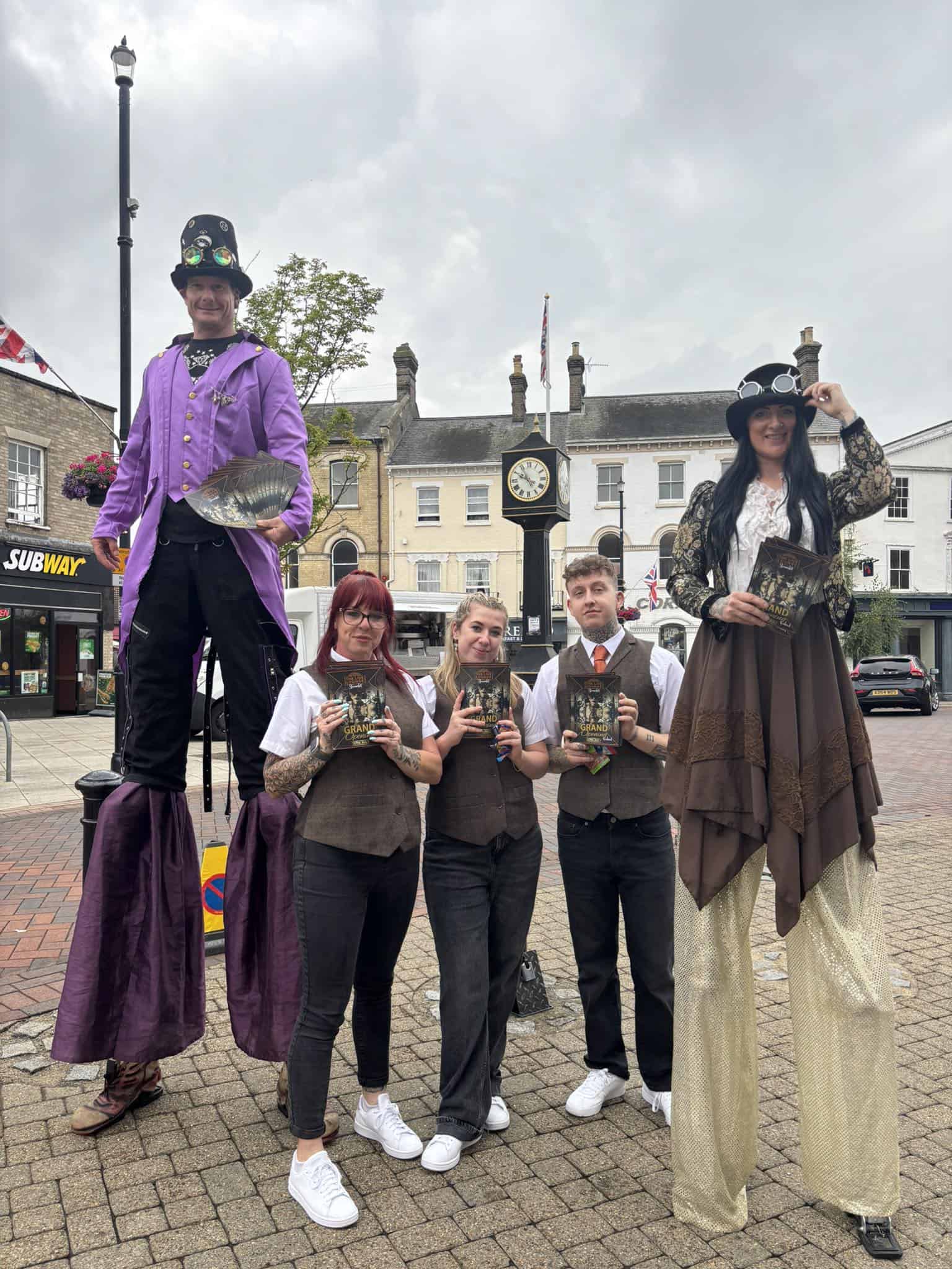 Vibrant steampunk performers in vintage attire with costumes, stilts, and accessories, entertaining visitors in front of The Live Lounge, showcasing live entertainment and creative street performances.