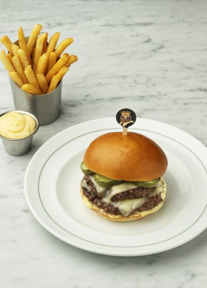 A double-patty cheeseburger with pickles on a brioche bun, served on a white plate next to a stainless steel cup of golden french fries and a small cup of dipping sauce, all on a white marble countertop.