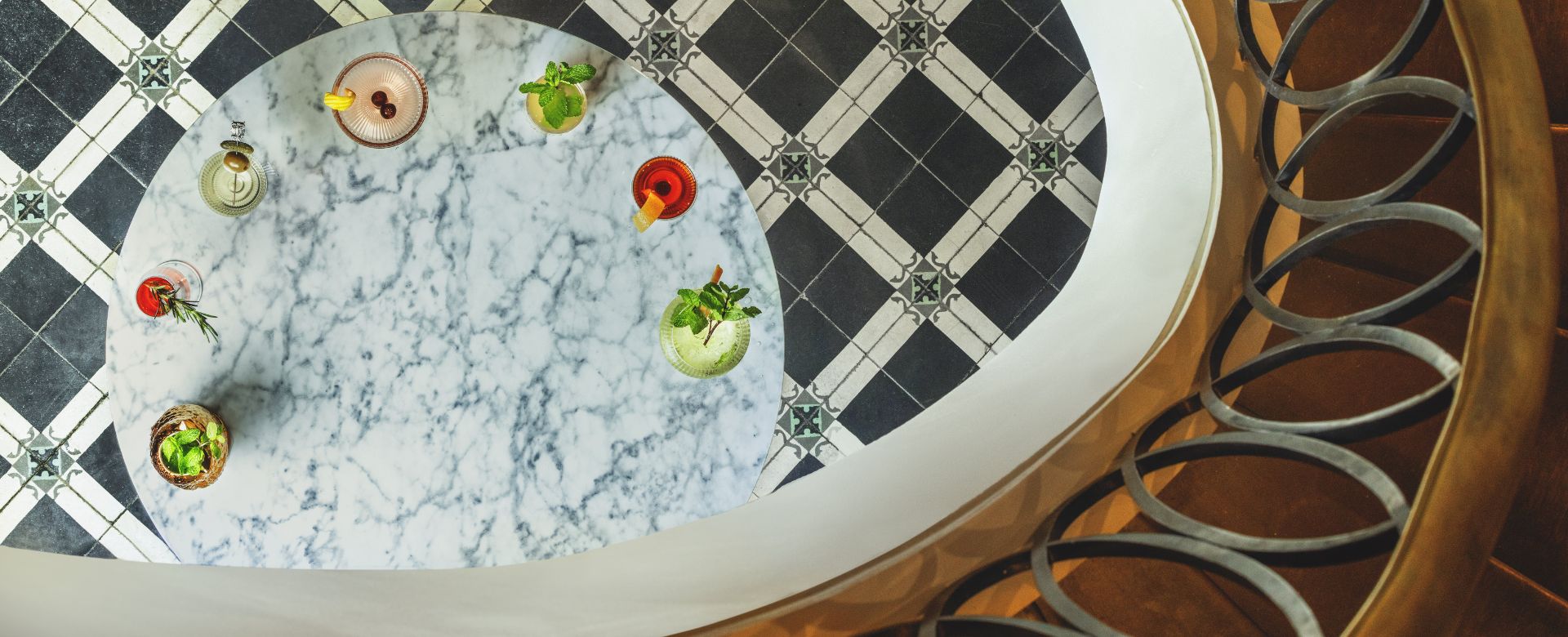 An artistic overhead shot looking down a curved, brass-railed spiral staircase to a small, round marble table on a black and white tiled floor, with a blue chair visible underneath the arch.