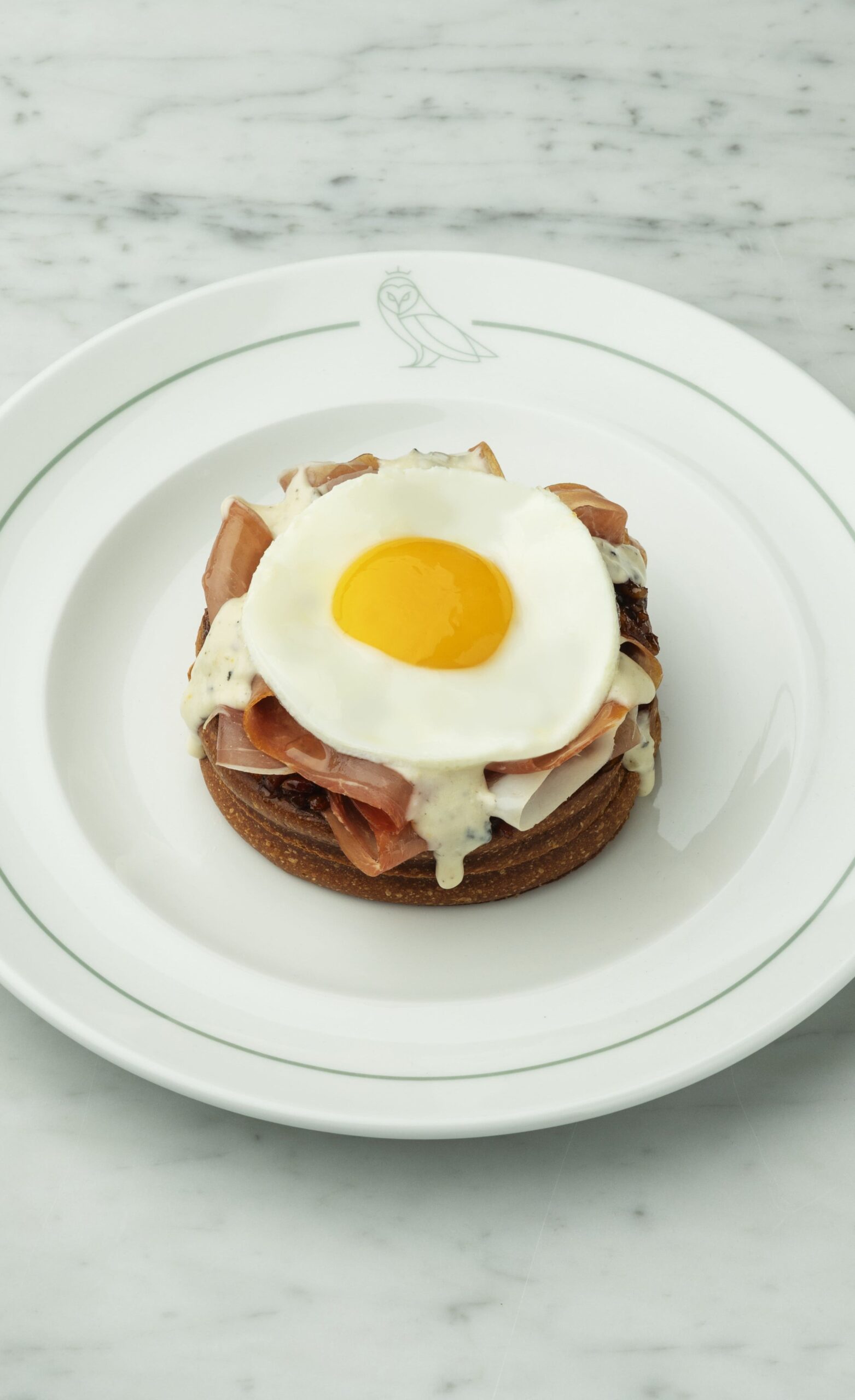 A gourmet open-face sandwich or toast topped with thin slices of prosciutto or ham, a creamy white sauce, and a perfectly cooked sunny-side-up egg, served on a white plate with an owl logo on a marble background.