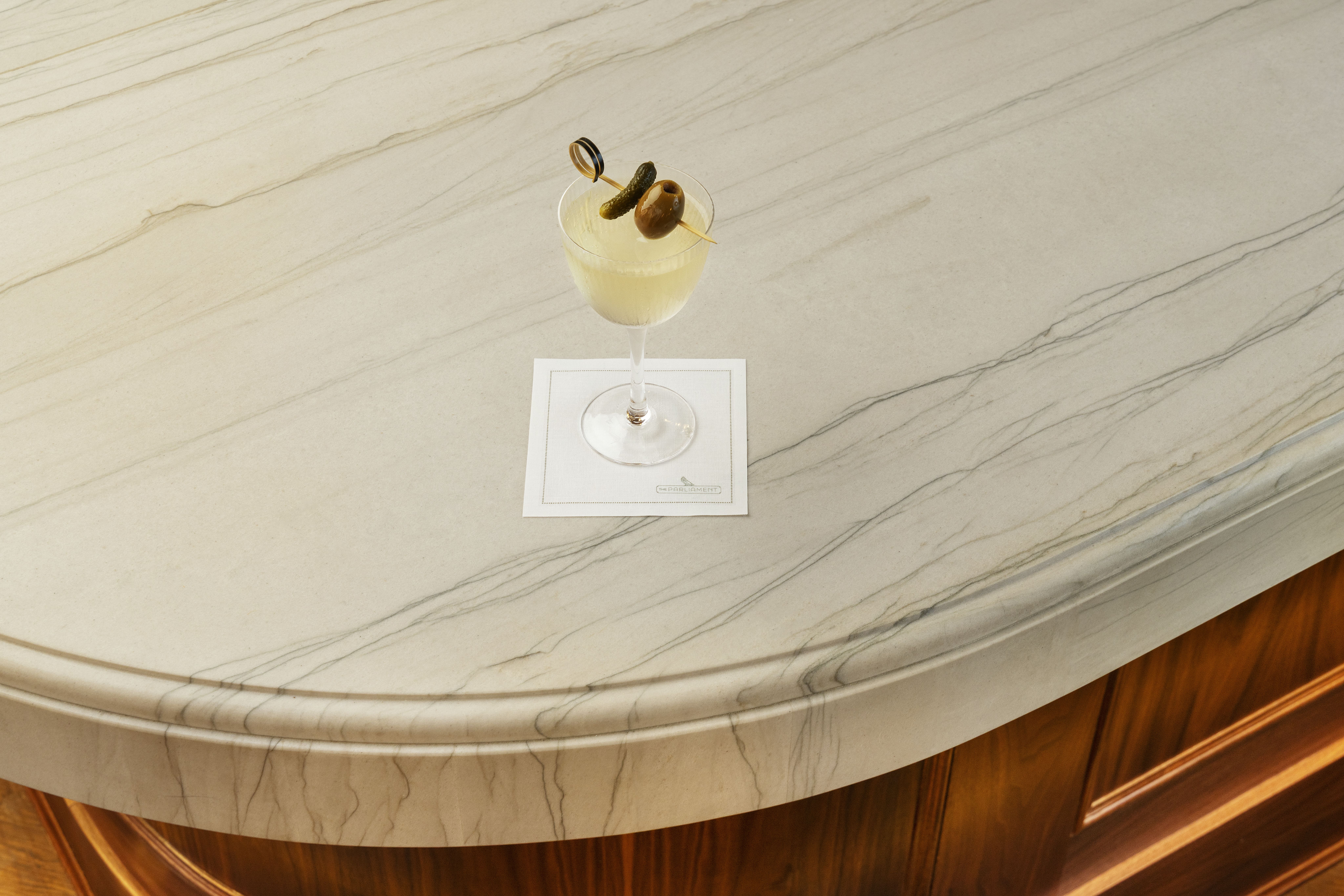 A pale yellow or clear martini cocktail in a stemmed coupe glass, garnished with two olives skewered on a pick, sitting on a white cocktail napkin on a smooth, veined marble bar top with a dark wood base.