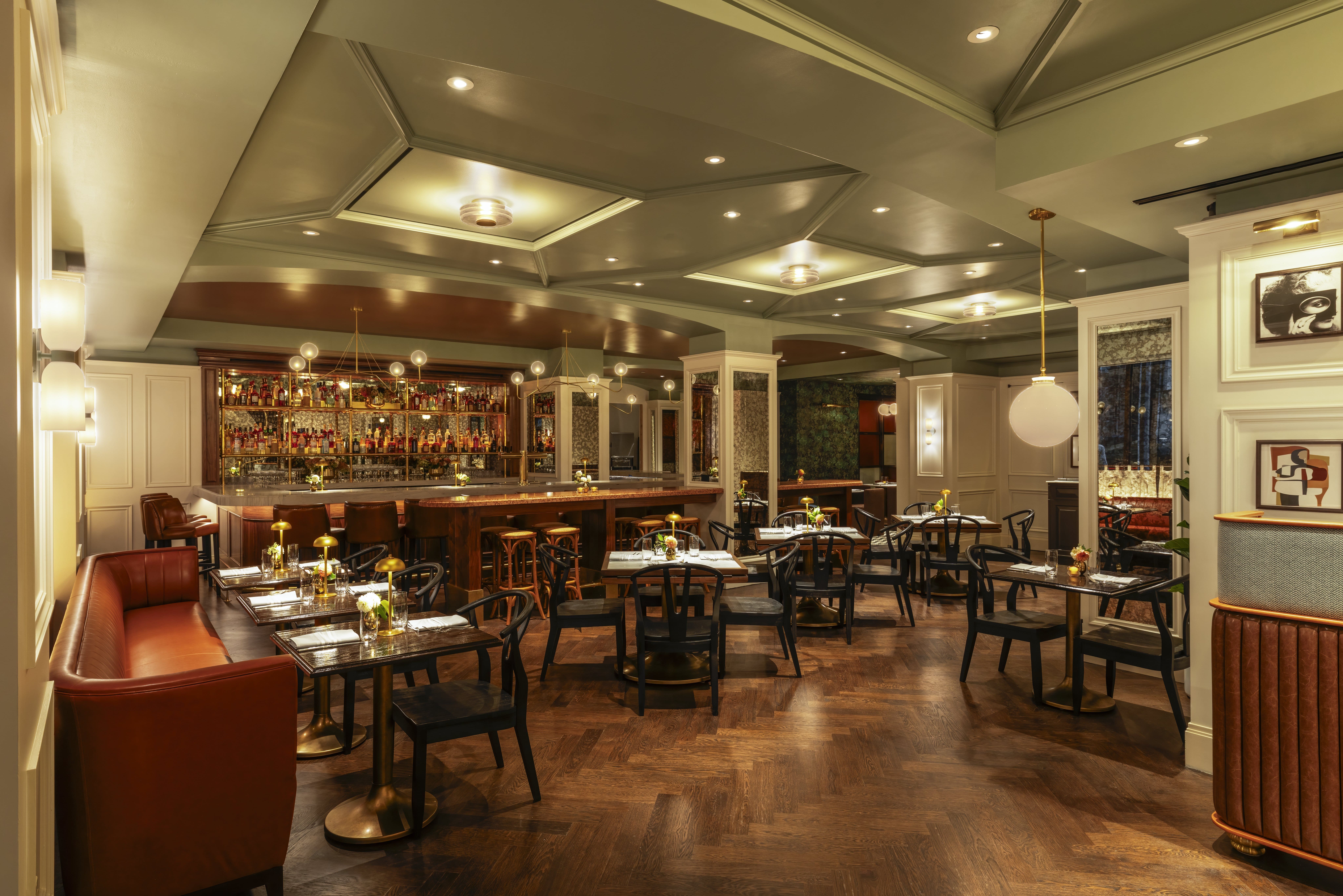 Parliament Main Dining and Bar A spacious, sophisticated restaurant interior with a dark wood chevron floor, featuring a long, red leather banquette along the left, multiple small tables with black chairs, and a well-lit, fully stocked bar in the background.