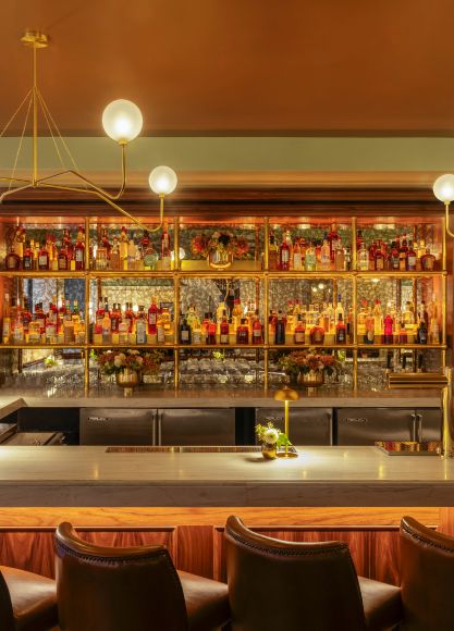 The interior of a sophisticated restaurant dining area and bar, featuring dark wood floors, black tables and chairs, a long red leather banquette, and a fully stocked back-lit bar.