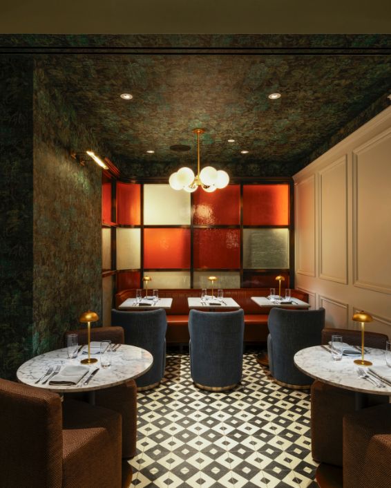 A private or intimate dining booth area with a colorful patterned ceiling, white paneled walls, a red leather banquette, and two marble-top tables with plush seating in the foreground.