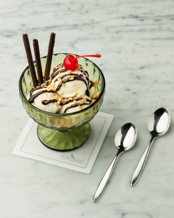 A classic ice cream sundae in a green glass goblet on a white marble surface, topped with chocolate sauce, chopped nuts, two chocolate sticks, and a bright red cherry. Two silver spoons rest beside it.