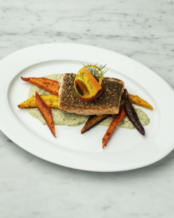 An elegantly plated entree featuring a piece of crispy, grilled branzino on an oval white plate, served over a light green sauce and surrounded by roasted orange and purple carrots, garnished with a slice of citrus.