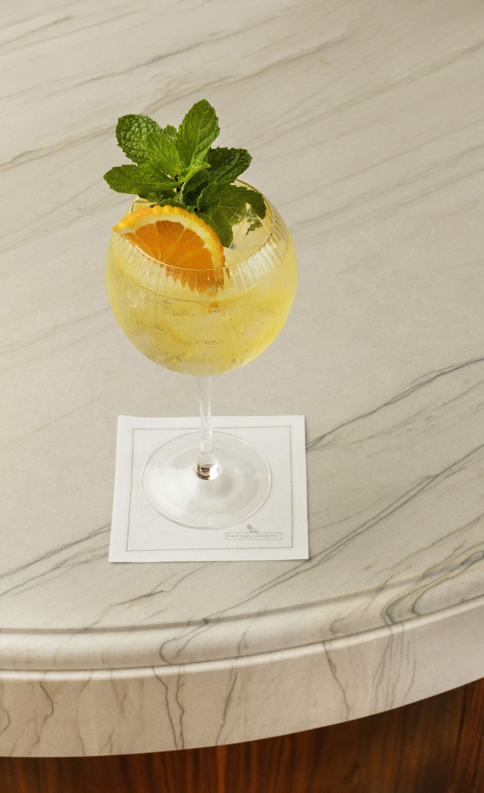 A sparkling, pale yellow cocktail served in a stemmed wine glass, garnished with a sprig of mint and a slice of orange, sitting on a white napkin on a curved marble bar top.