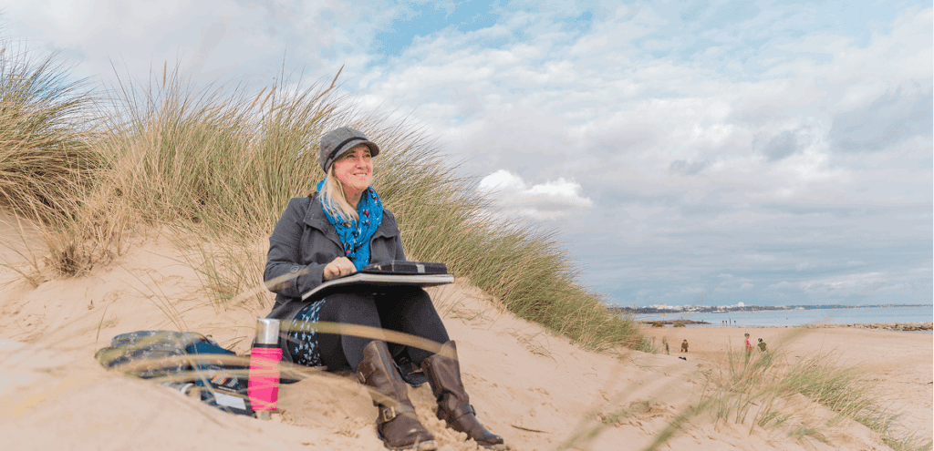 Artist, Nina Squire, sitting on a beach with a sketchbook