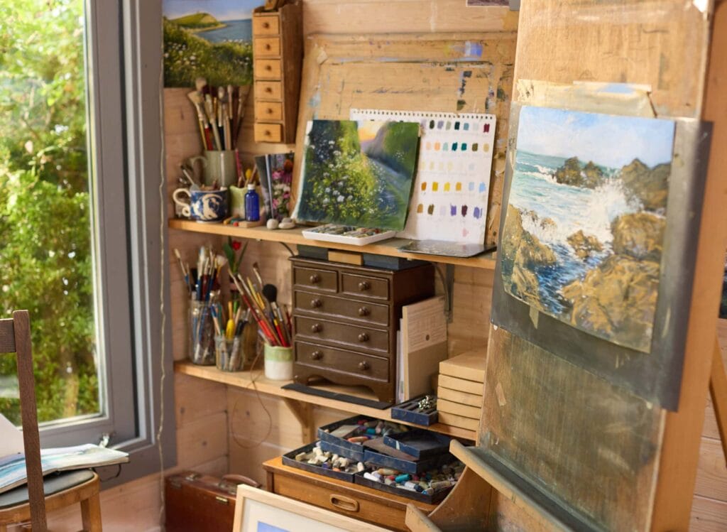 Artist's studio with paintings, brushes, and art supplies by a window.