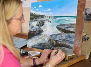Nina Squire creating a detailed seascape painting of rocks and waves.