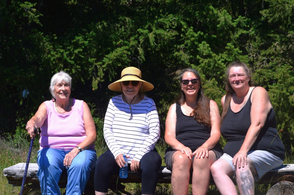 Four women sit on a wooden bench outdoors in sunlight, with trees in the background. One woman wears a sunhat, and all are smiling at the camera.