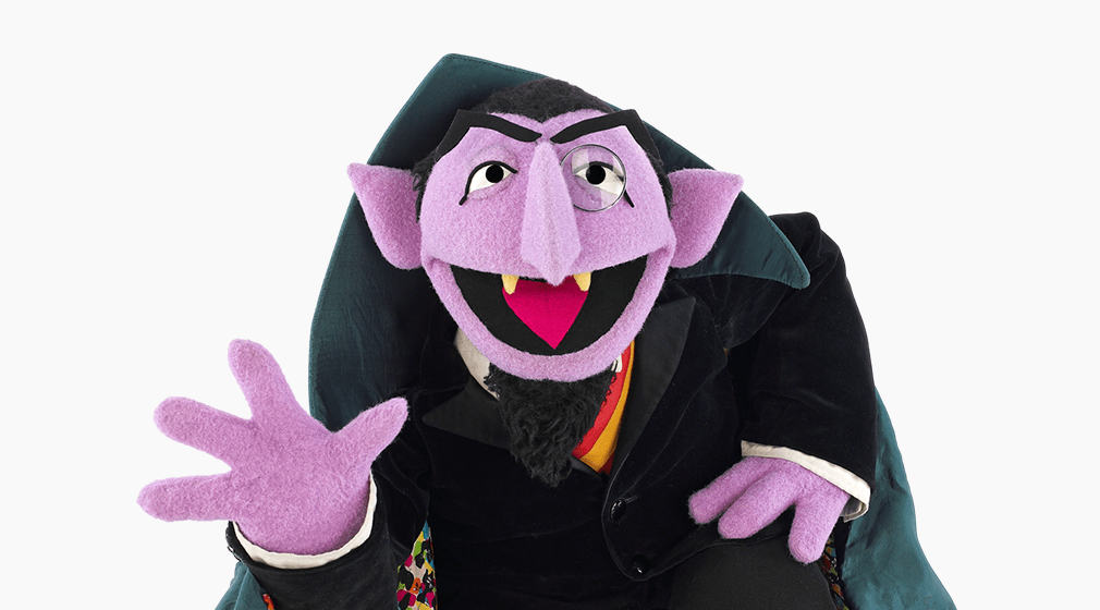 A purple puppet character with fangs, wearing a black suit and a red bow tie, sitting on a chair.