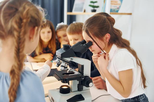 A group of children in a classroom gather around a table while one girl looks through a microscope, with books and a small plant in the background.