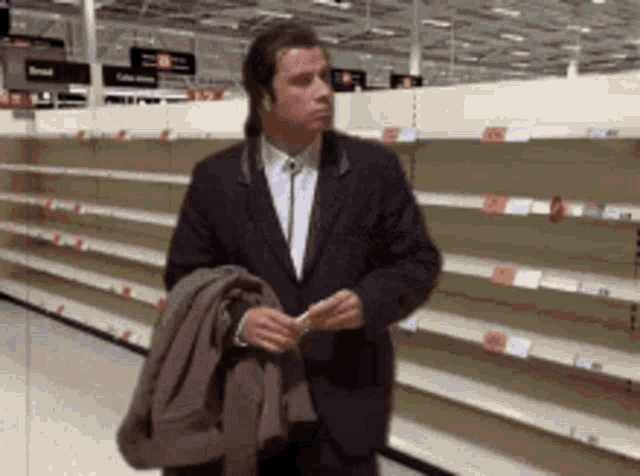 A man in a coat stands in a nearly empty grocery store aisle, looking around at the bare shelves.