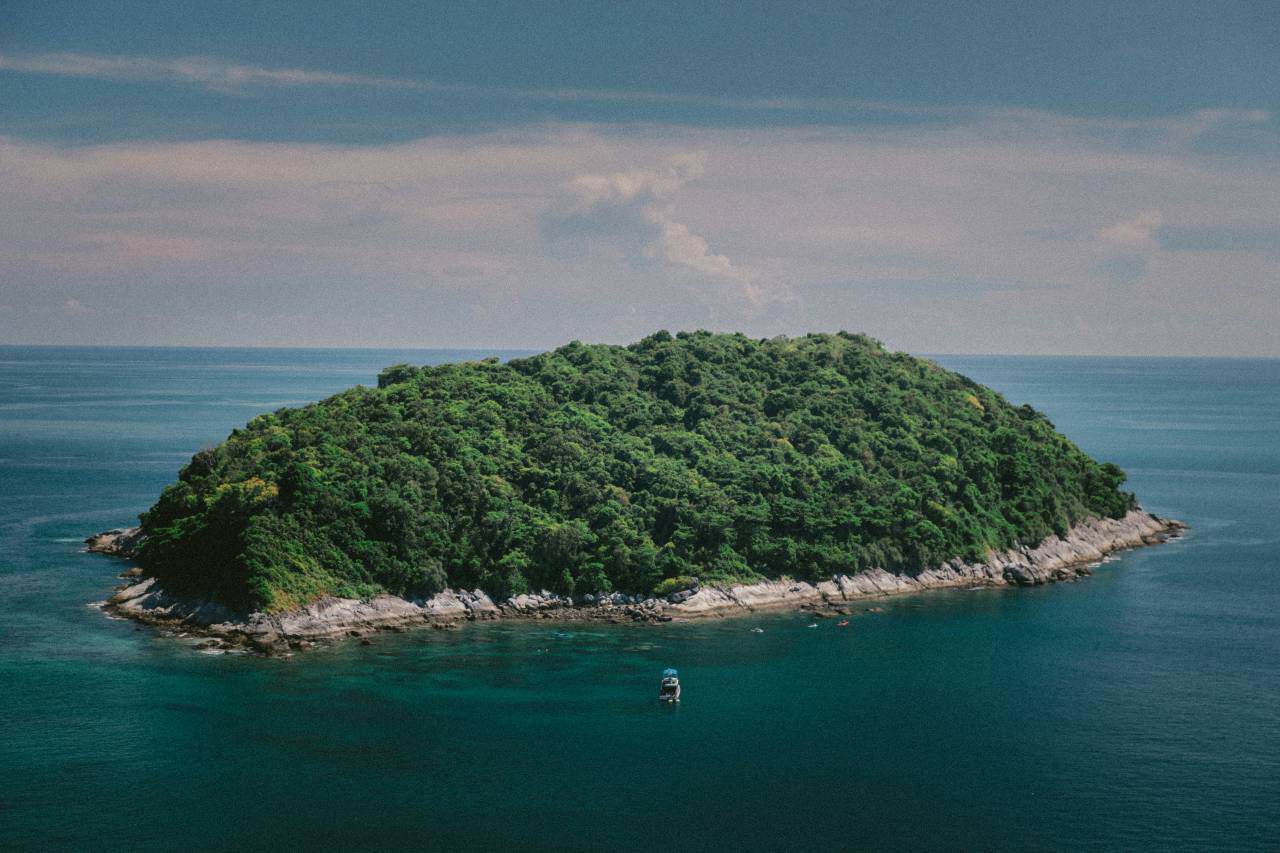A small, densely forested island surrounded by clear blue water, with a single boat anchored nearby.