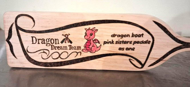 Wooden paddle with "Dragon Dream Team" and "dragon boat pink sisters paddle as one" engraved, featuring a small pink cartoon dragon illustration.