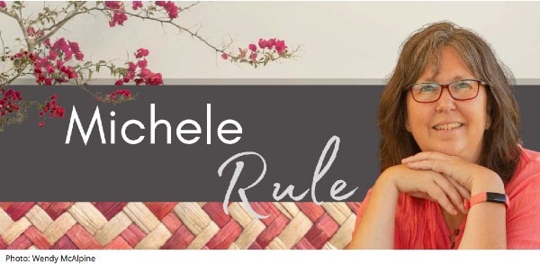 A woman with glasses and gray hair sits smiling beside woven decor and flowers. Text reads “Michele Rule.” Photo credit: Wendy McAlpine.