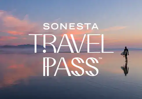 Image of a person standing in shallow water reflecting the sky with the Sonesta Travel Pass Logo overlaid.