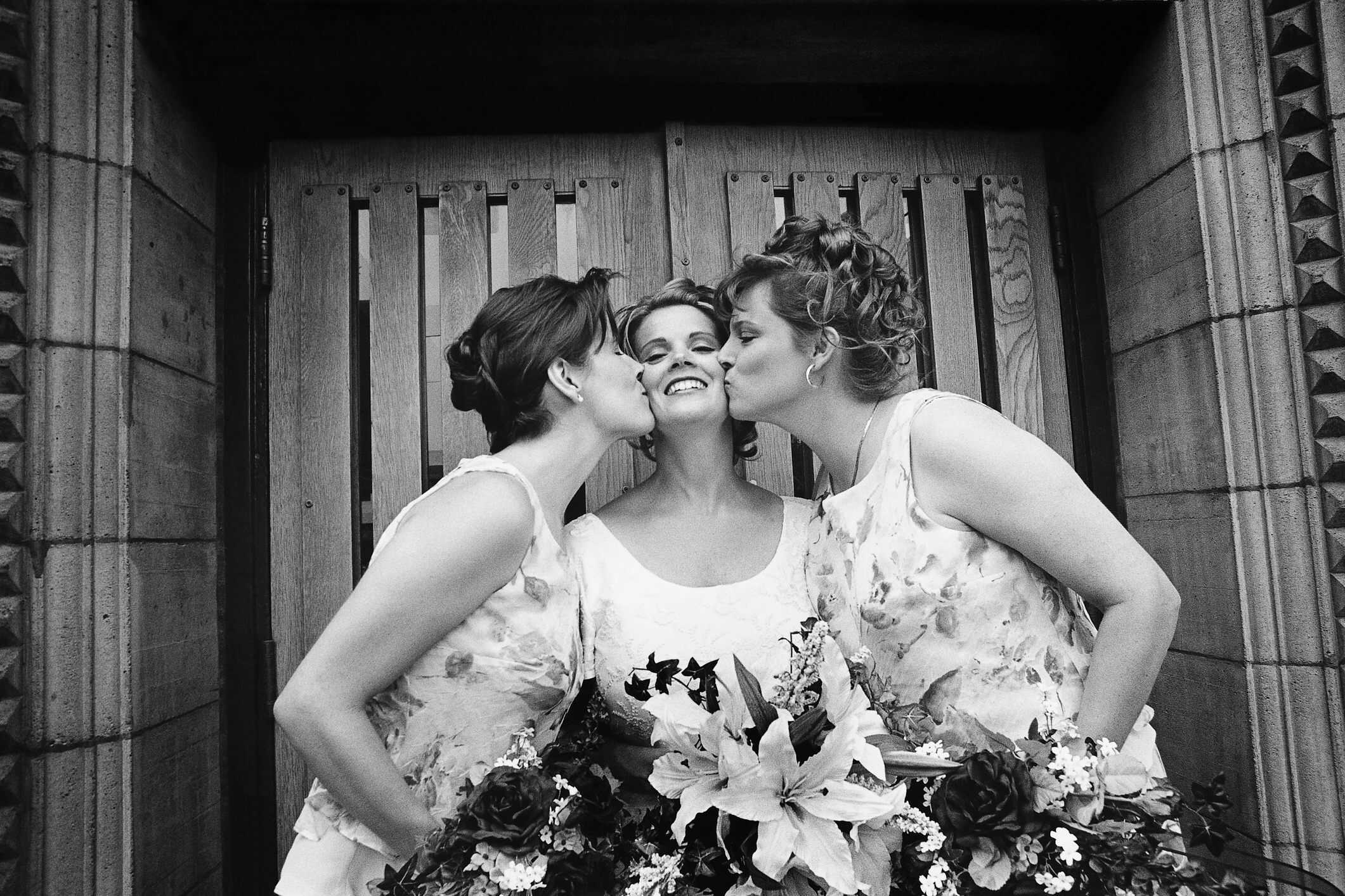 Black and white photo of a smiling bride being kissed on both cheeks by two bridesmaids holding floral bouquets