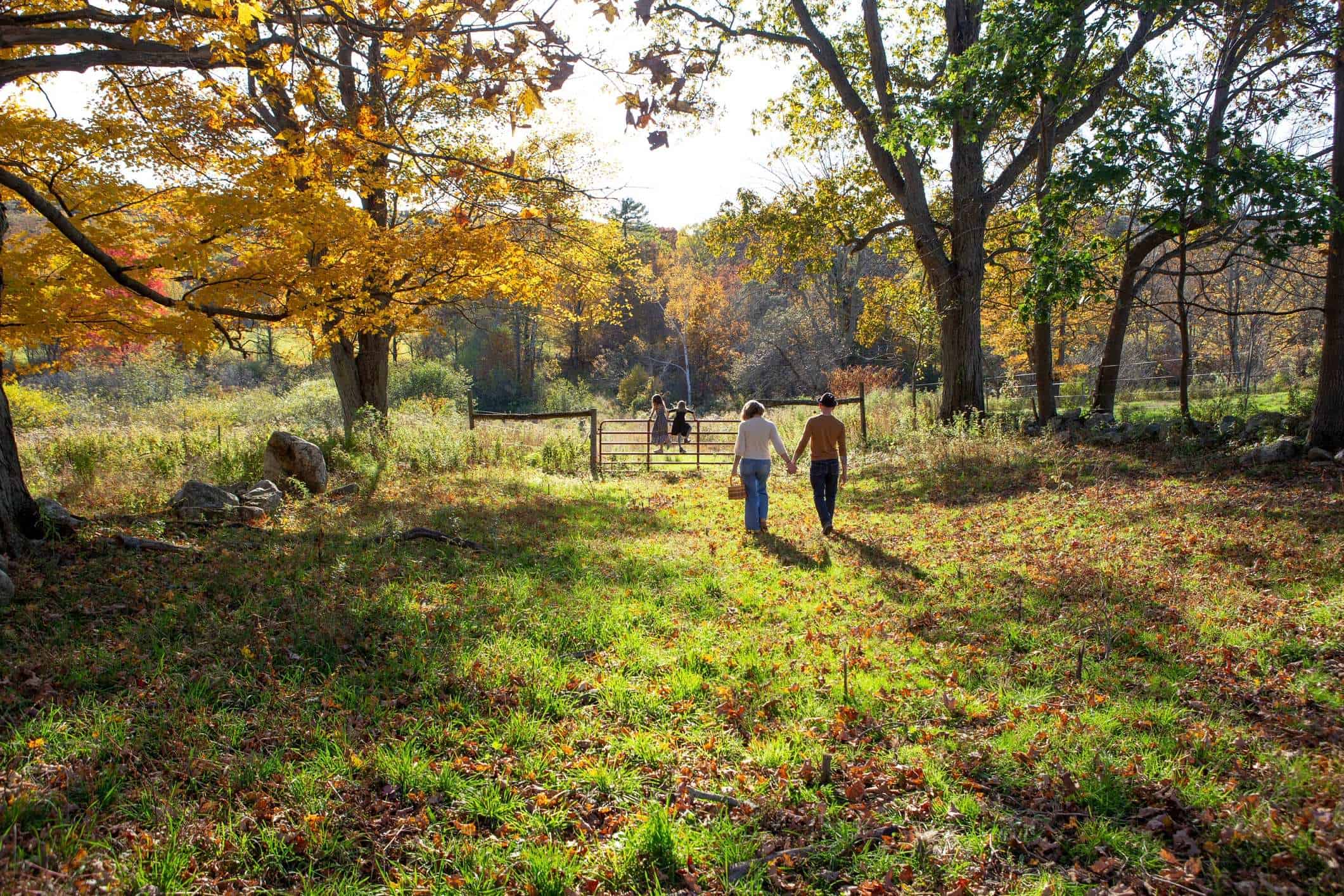 Family walking hand-in-hand through an autumn countryside meadow toward a farm gate