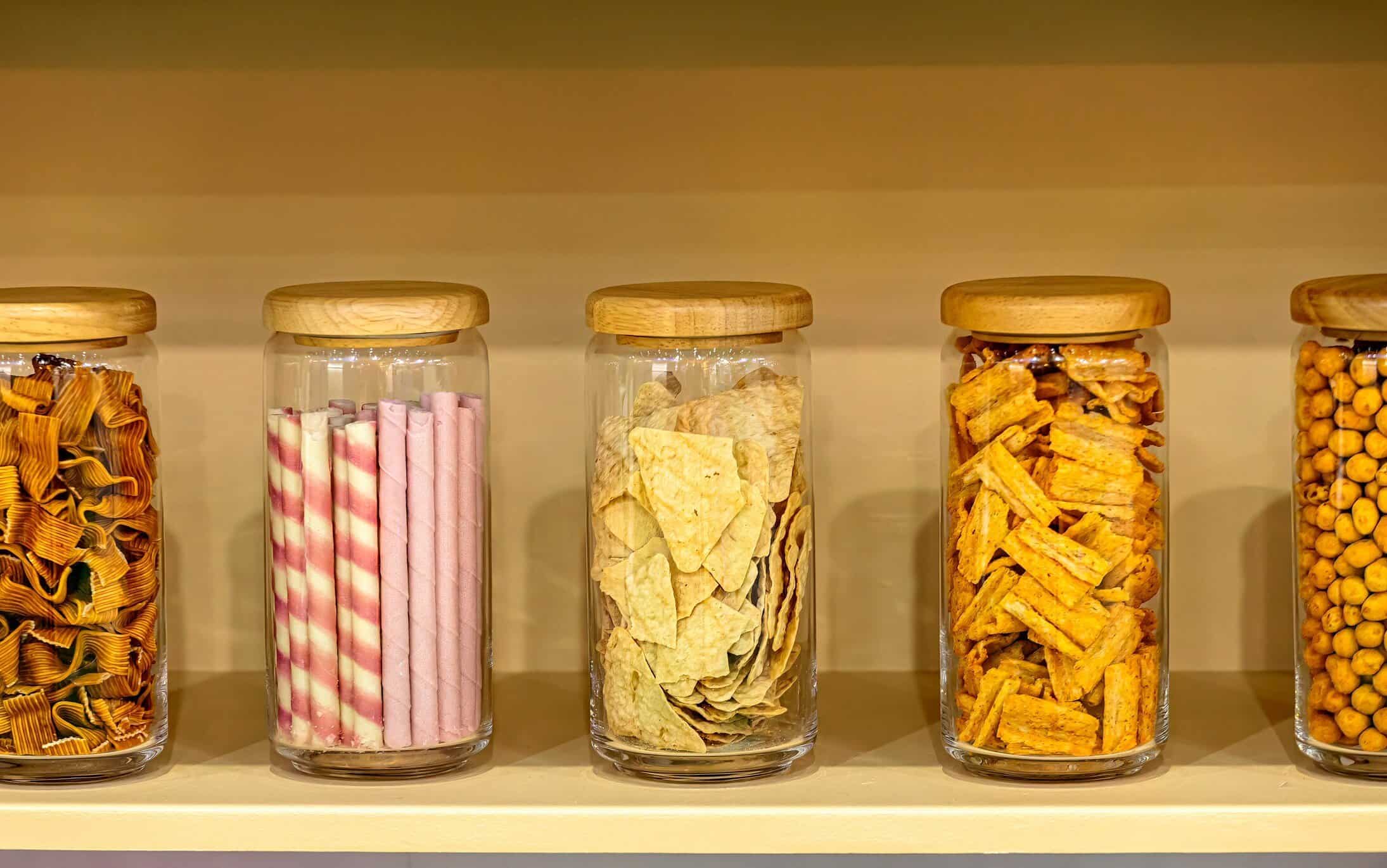 Row of glass jars with wooden lids containing assorted snacks and wafer rolls on a shelf